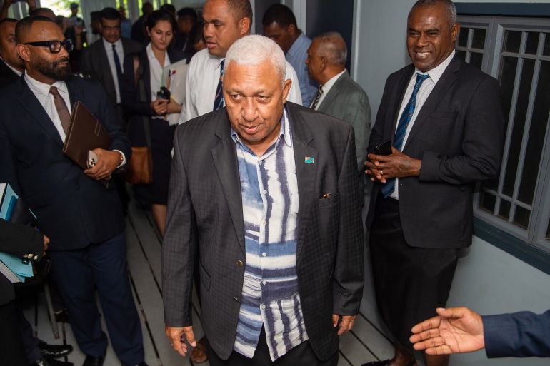 Fiji's former prime minister Frank Bainimarama. He is wearing a blue and white patterned shirt with a jacket. His hair is white. He is arriving in court. There are other people nearby.